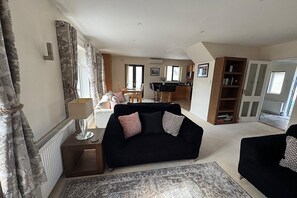 Ein Wohnzimmer mit einer schwarzen Couch, einem Couchtisch und einem Teppich mit Muster. Es gibt einen Essbereich mit Tisch und Stühlen, und im Hintergrund eine Küche.