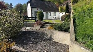 Uma casa branca com telhado preto, cercada por um jardim com caminhos de cascalho e diversas plantas.