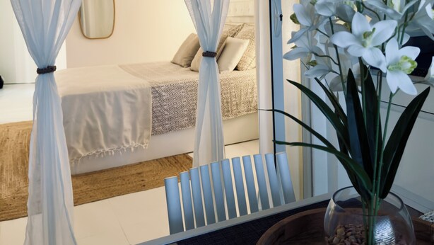 Une chambre à coucher avec un lit, une table de chevet ornée d’un vase de fleurs, des rideaux légers et une vue sur l’extérieur.