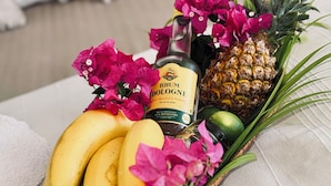 Un panier en osier contenant des bananes, une bouteille de rhum et un ananas, le tout posé sur un lit.