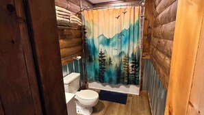 A bathroom with wooden walls, a toilet, a shower curtain with a nature scene, and a wooden floor.
