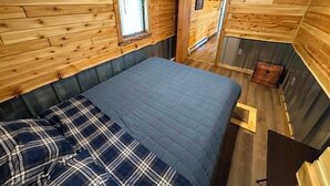 A wooden cabin interior with a bed, a window with blinds, and a door leading to another room.