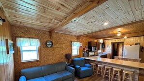 A cabin interior with a wooden ceiling, a sofa, a bar area with stools, and a kitchen.