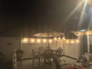 Un espace extérieur aménagé en terrasse, avec une table ronde et des chaises, le tout illuminé par des guirlandes lumineuses.
