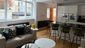 A modern living room with a sofa, two coffee tables, and a kitchen area with bar stools. - West End Elegance - by Out of Office Lifestyle (London)