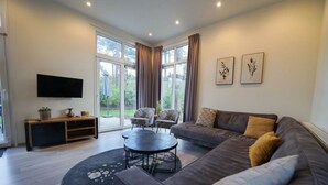 A modern living room with a sectional sofa, a round coffee table, a flat-screen TV, and a sliding glass door leading to a garden. - Holiday Home in Otterlo With Terrace (Otterlo)