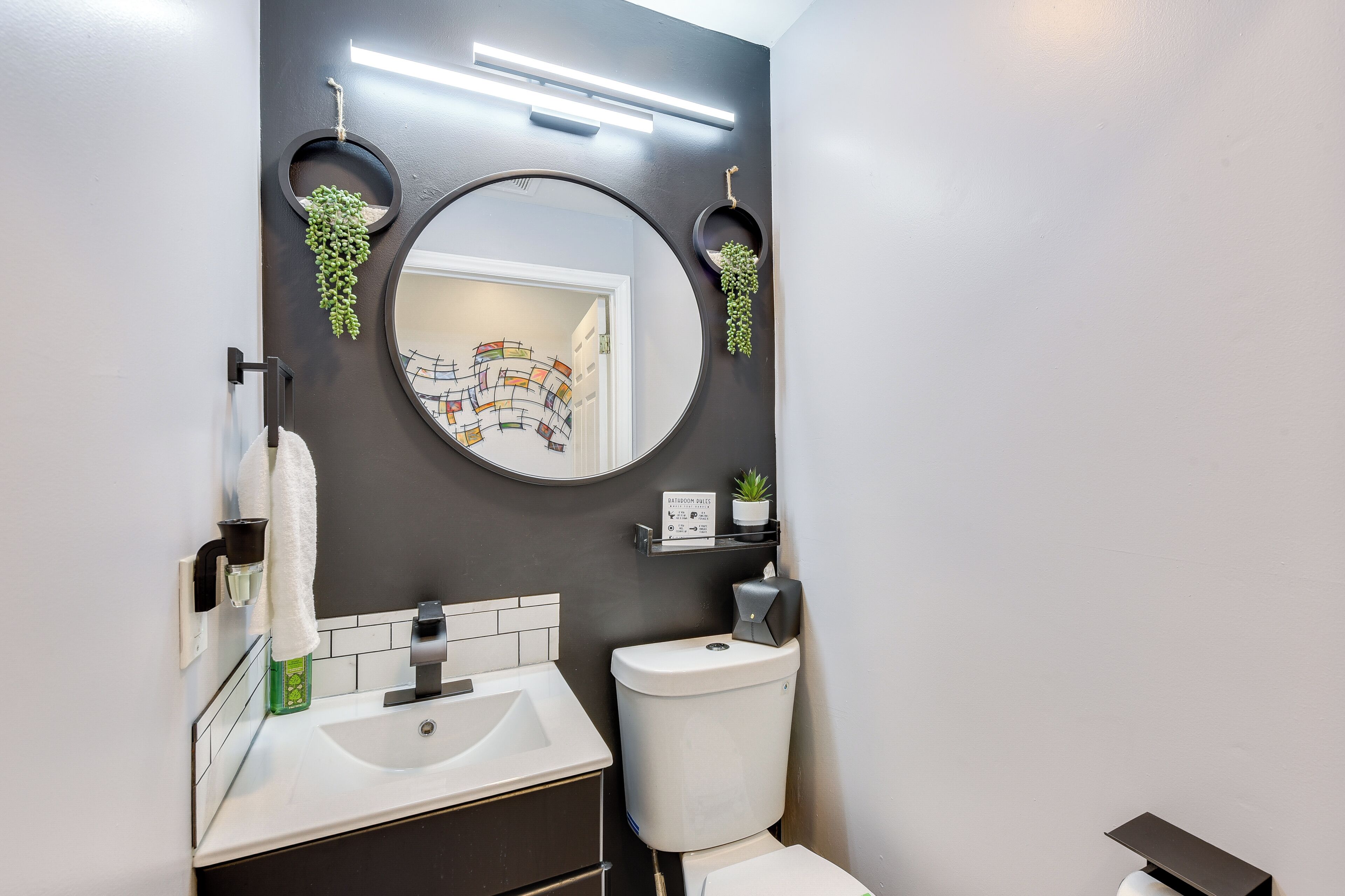Ein modernes Badezimmer mit einem weißen Waschbecken, einer Toilette, einem runden Spiegel und zwei Regalen an der Wand mit Pflanzen.