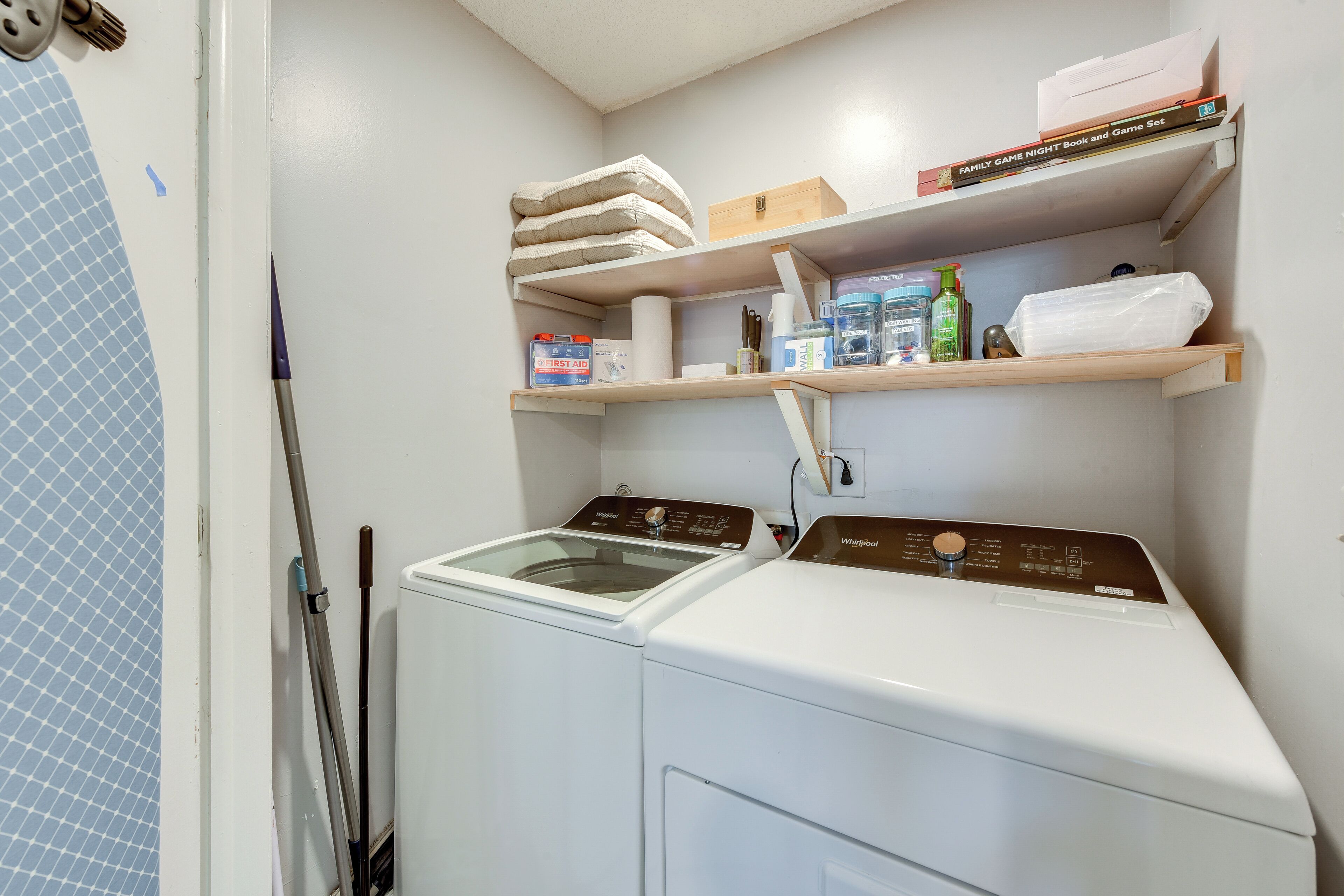 A laundry area with a front-loading washer and dryer, shelves with books and toiletries, and a mop leaning against the wall.