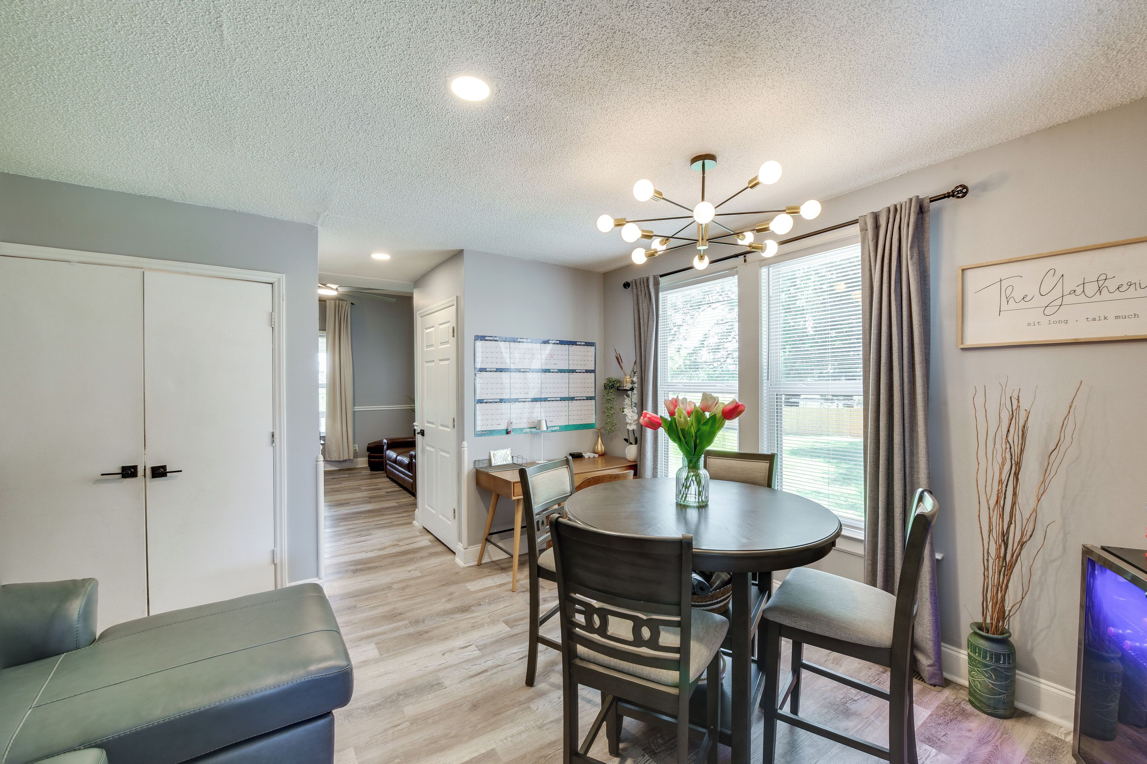 A dining area with a round table, chairs, a vase with flowers, a wall calendar, and a framed quote.