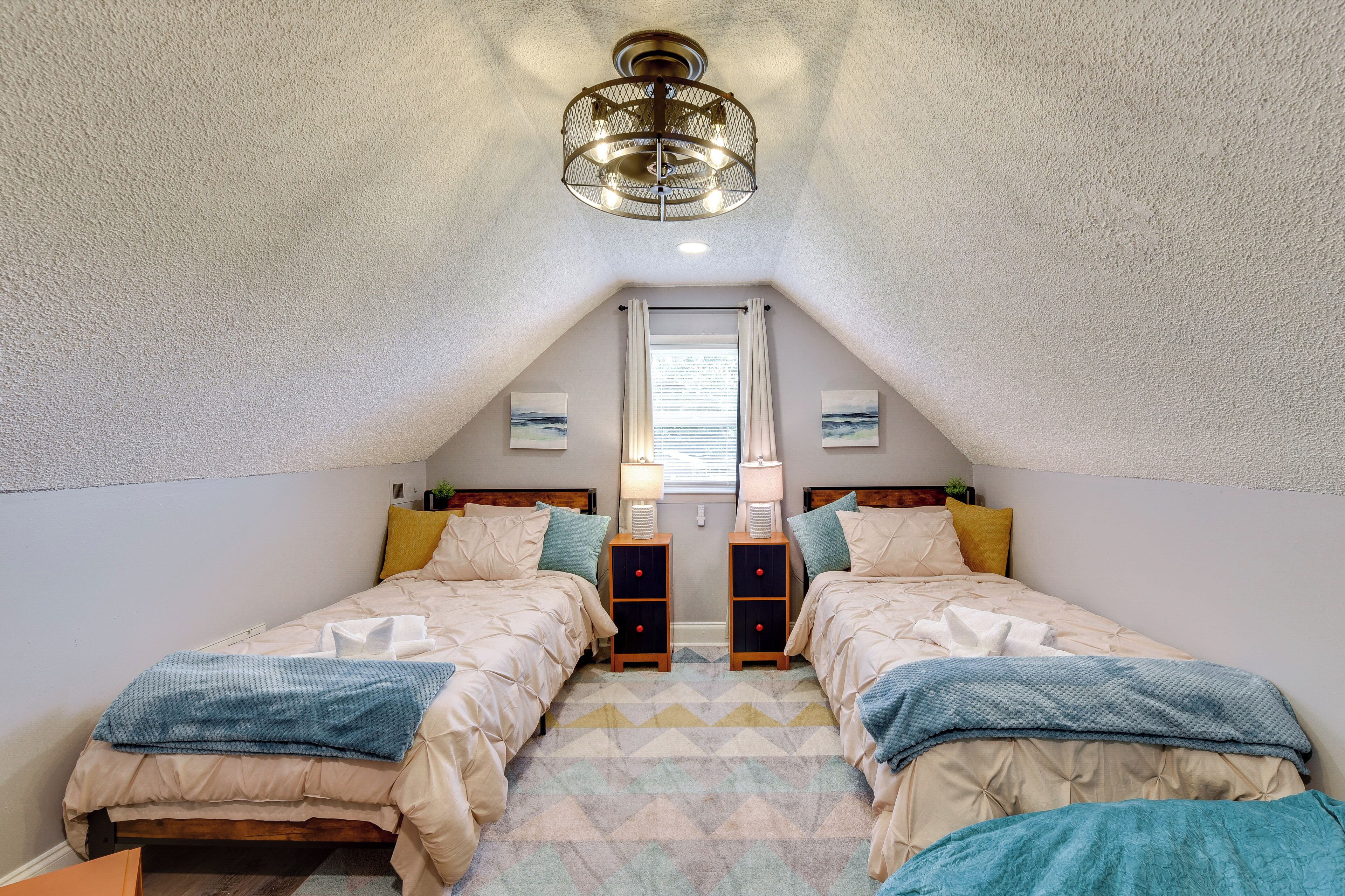 A bedroom with two beds, a chandelier, a window with curtains, and a geometric-patterned rug.