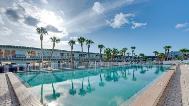 A swimming pool with clear water, surrounded by palm trees and a building with balconies. - Steps to Beach! Charming Indian Shores Condo (Indian Shores)