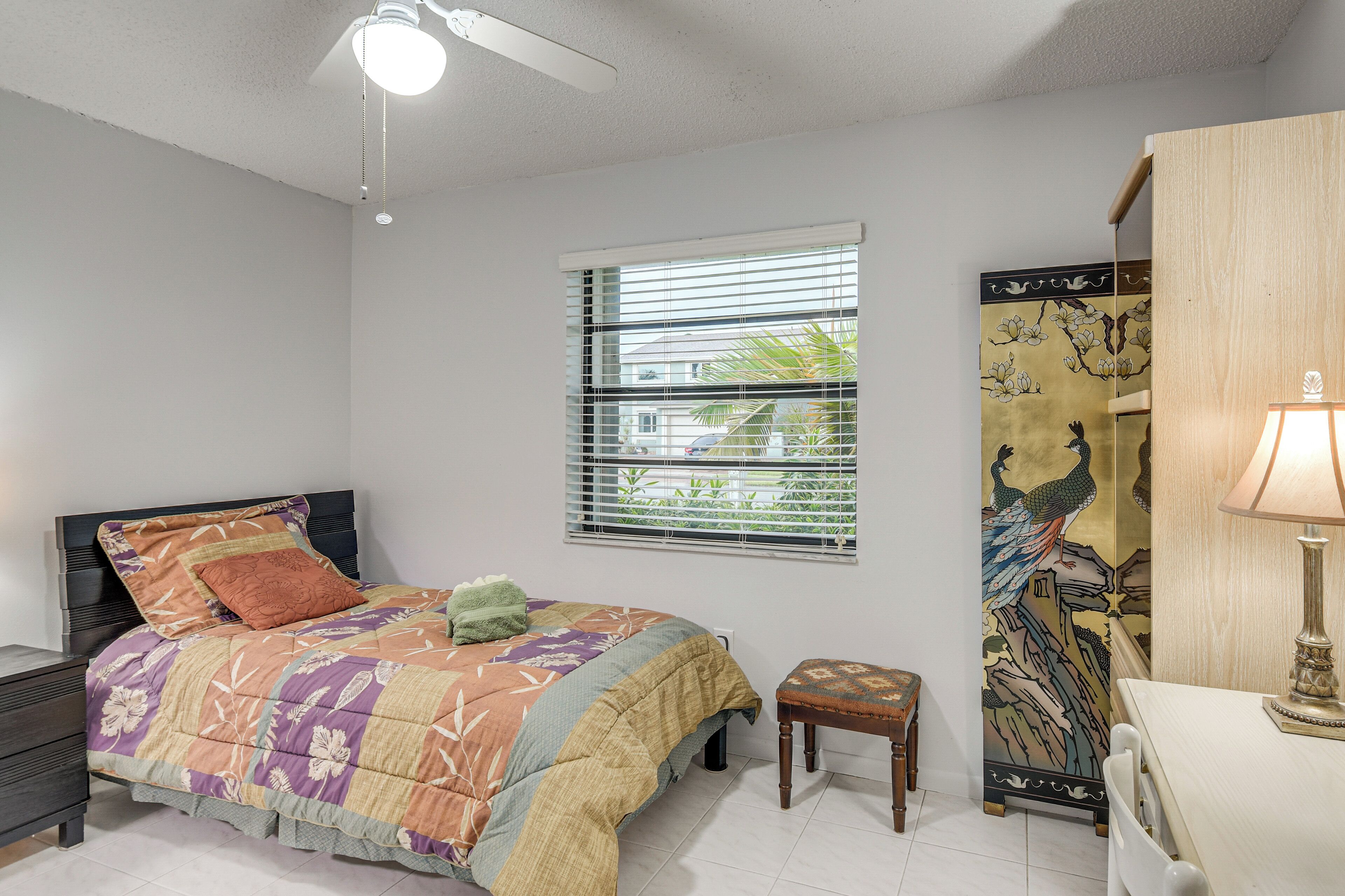 A bedroom with a bed, a nightstand, a window with blinds, a wall-mounted shelf, and a desk with a lamp.