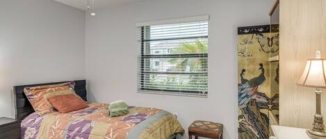 A bedroom with a bed, a nightstand, a window with blinds, a wall-mounted shelf, and a desk with a lamp.