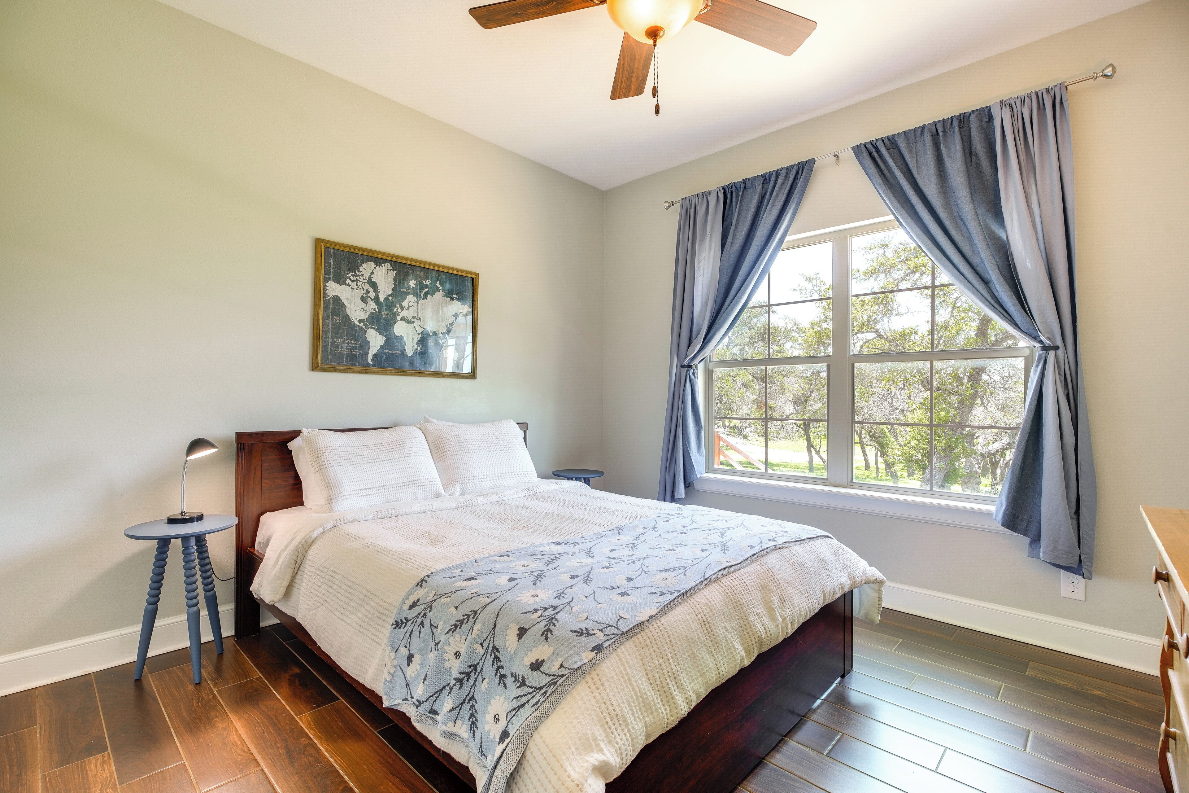 A bedroom with a bed, a side table, a lamp, a ceiling fan, a window with curtains, and a world map on the wall.