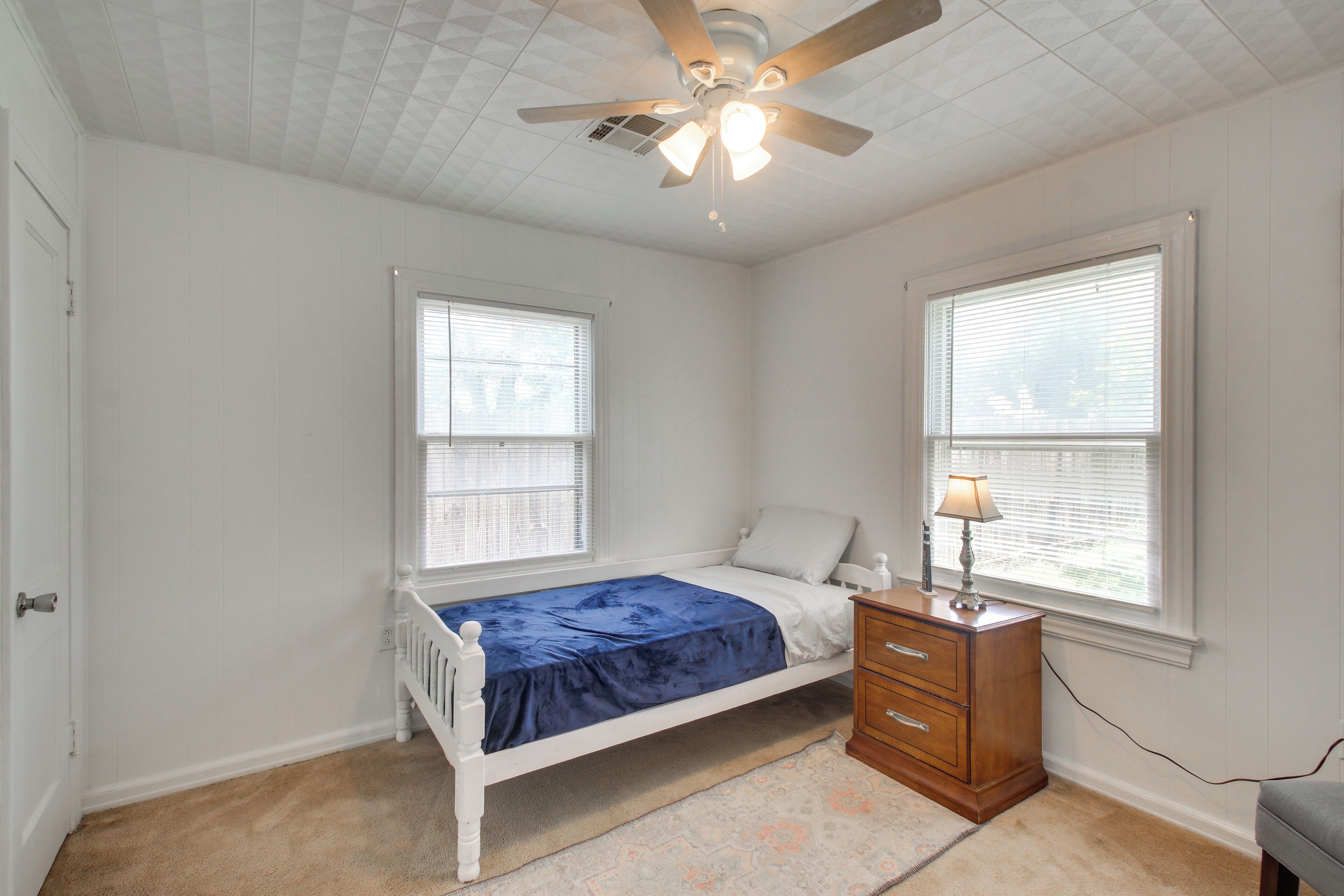 A bedroom with a single bed, a nightstand, a dresser, a ceiling fan, and two windows with blinds.