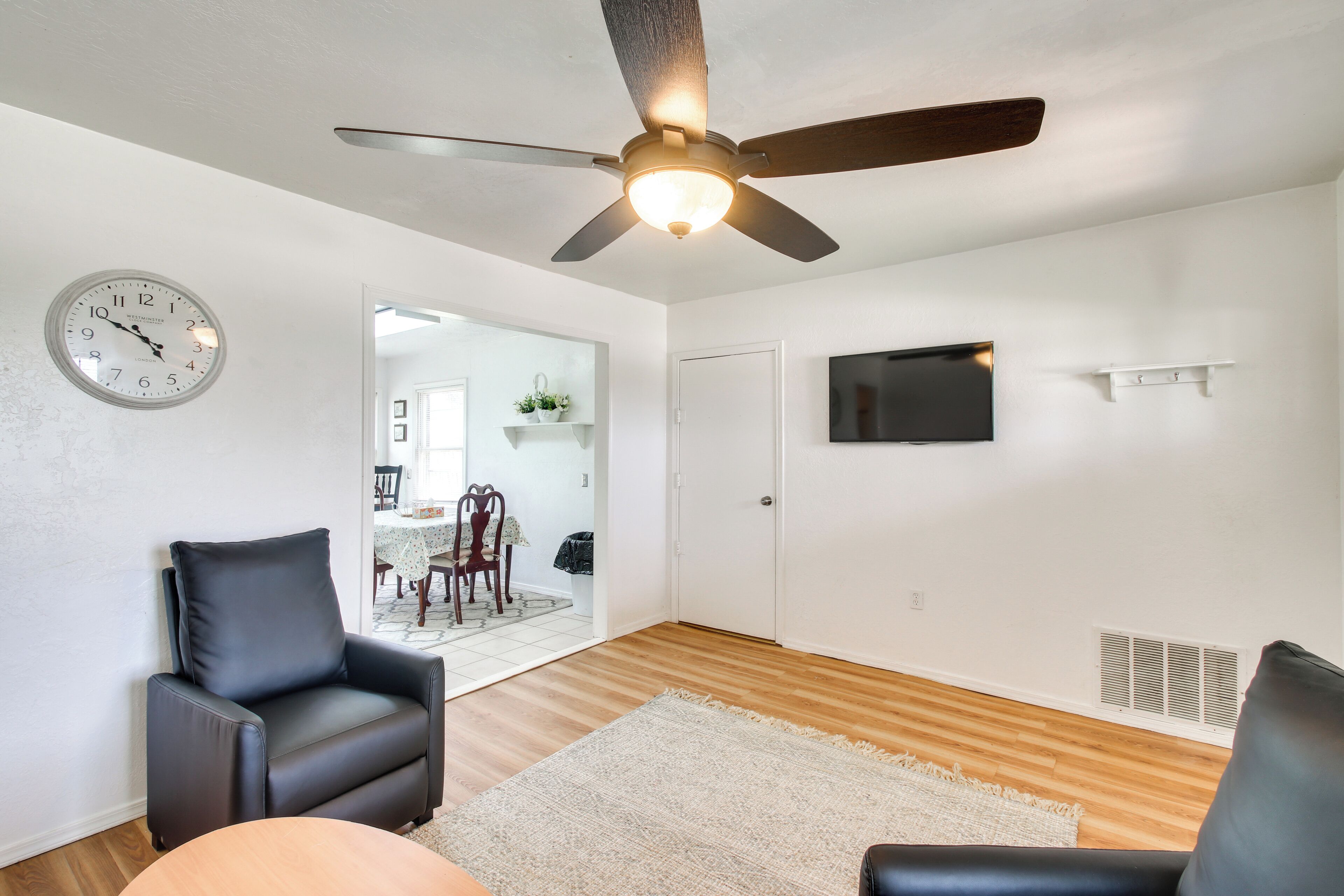 A living room with a ceiling fan, a clock, a flat-screen TV, and a dining area in the background.
