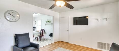 A living room with a ceiling fan, a clock, a flat-screen TV, and a dining area in the background.