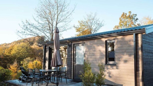 Una cabaña de madera moderna con un patio, mobiliario exterior y vistas al bosque circundante.