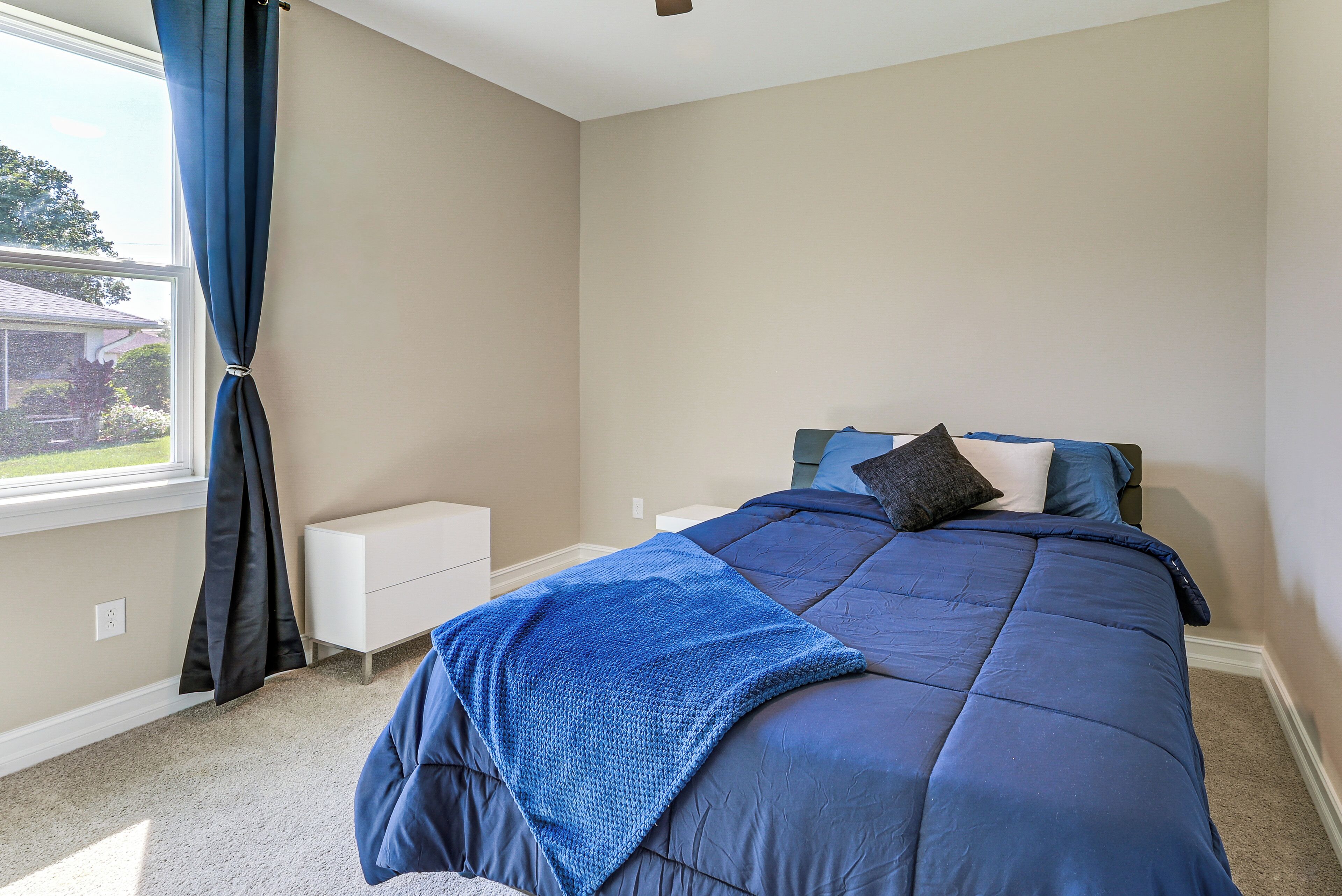 A bedroom with a bed, a nightstand, a window with curtains, and a view of a house outside.