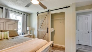 Une chambre à coucher comprenant un lit, une table de chevet, une commode, un ventilateur de plafond, une porte de placard coulissante de style rustique et une porte blanche.