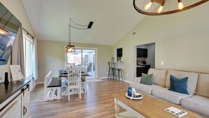 Un vaste espace de vie comprenant une table à manger en bois, des chaises blanches, un canapé avec des coussins et, au fond, une cuisine.