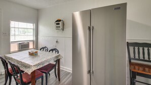 Ein Essbereich mit Tisch, bedeckt mit einer gemusterten Tischdecke, Holzstühle, einem Kühlschrank und einem Fenster mit Jalousien.