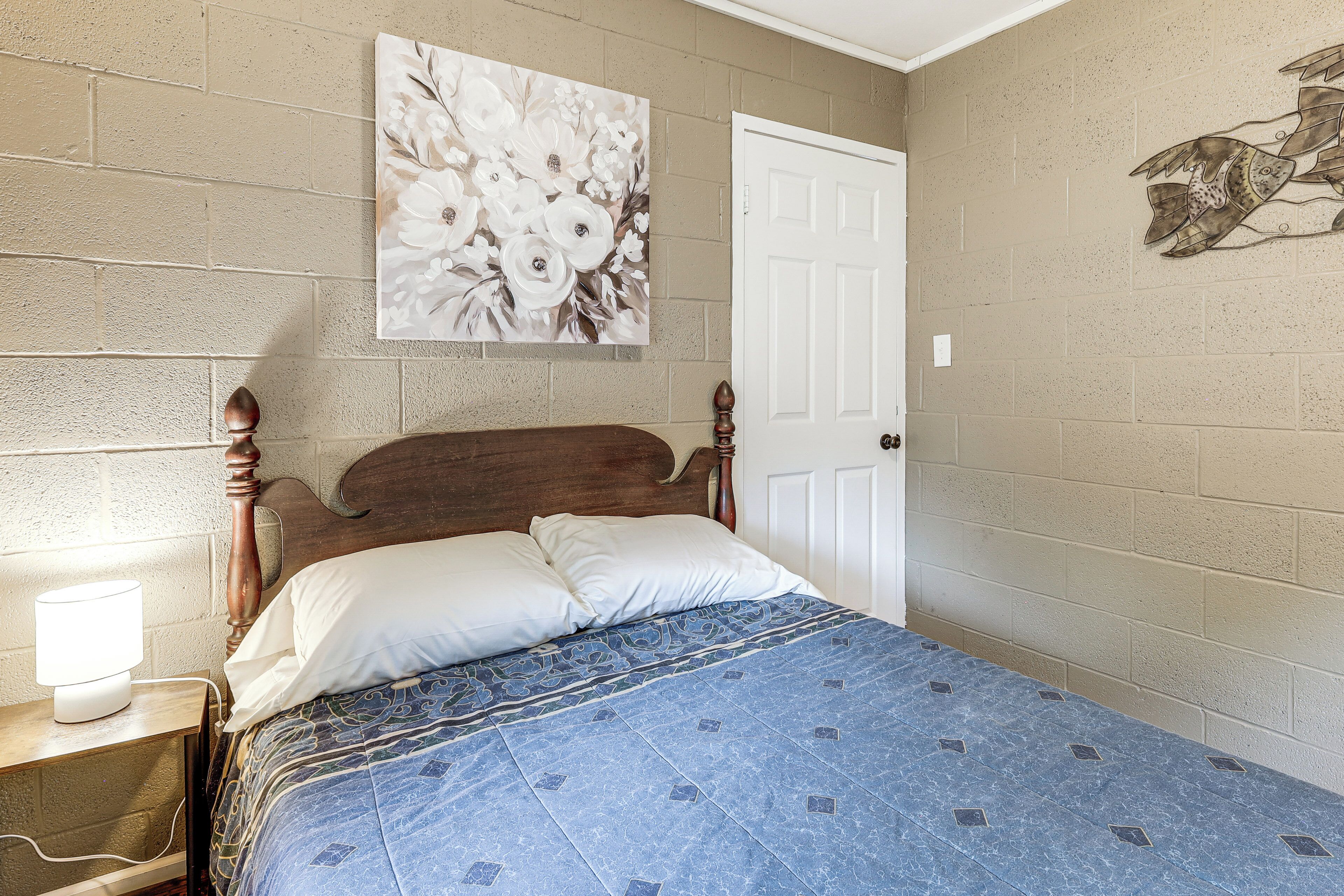 Een slaapkamer met een houten hoofdbord, een bed met een blauw beddegoed met een patroon, een witte deur en een bloemig kunstwerk aan de muur.