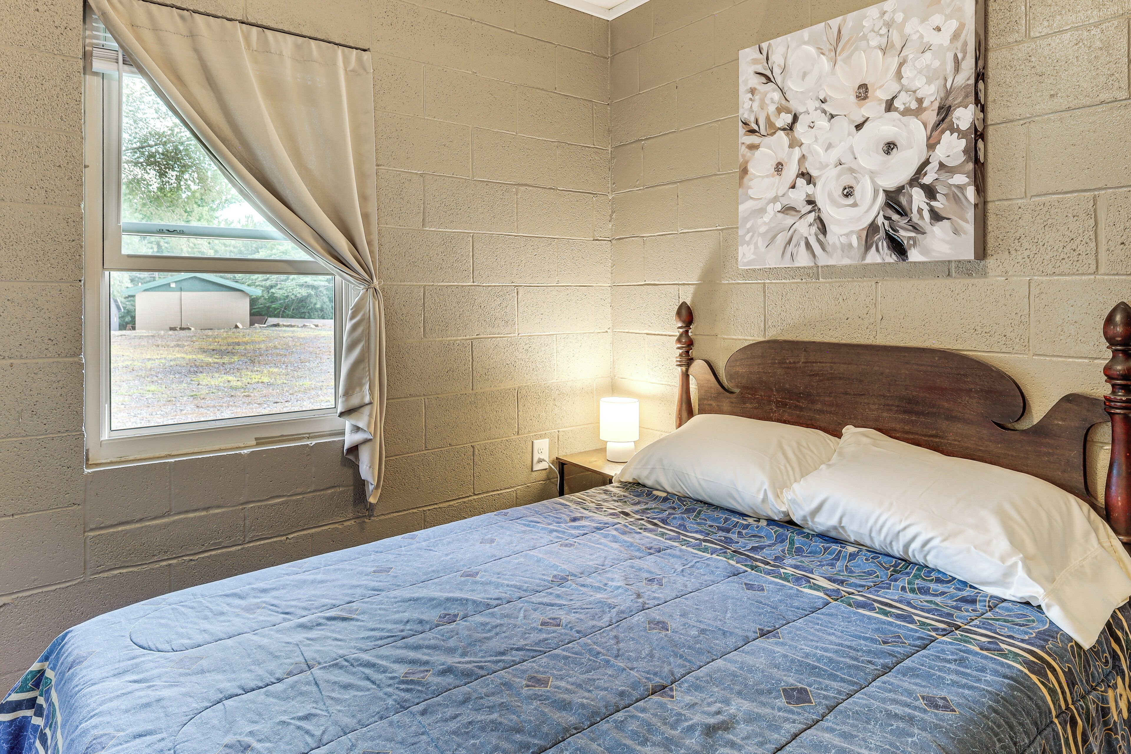 Un dormitorio con una cama, una ventana con cortinas, un cuadro en la pared y una mesita de noche con una lámpara.