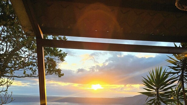 A patio with a view of a sunset over water, surrounded by greenery.