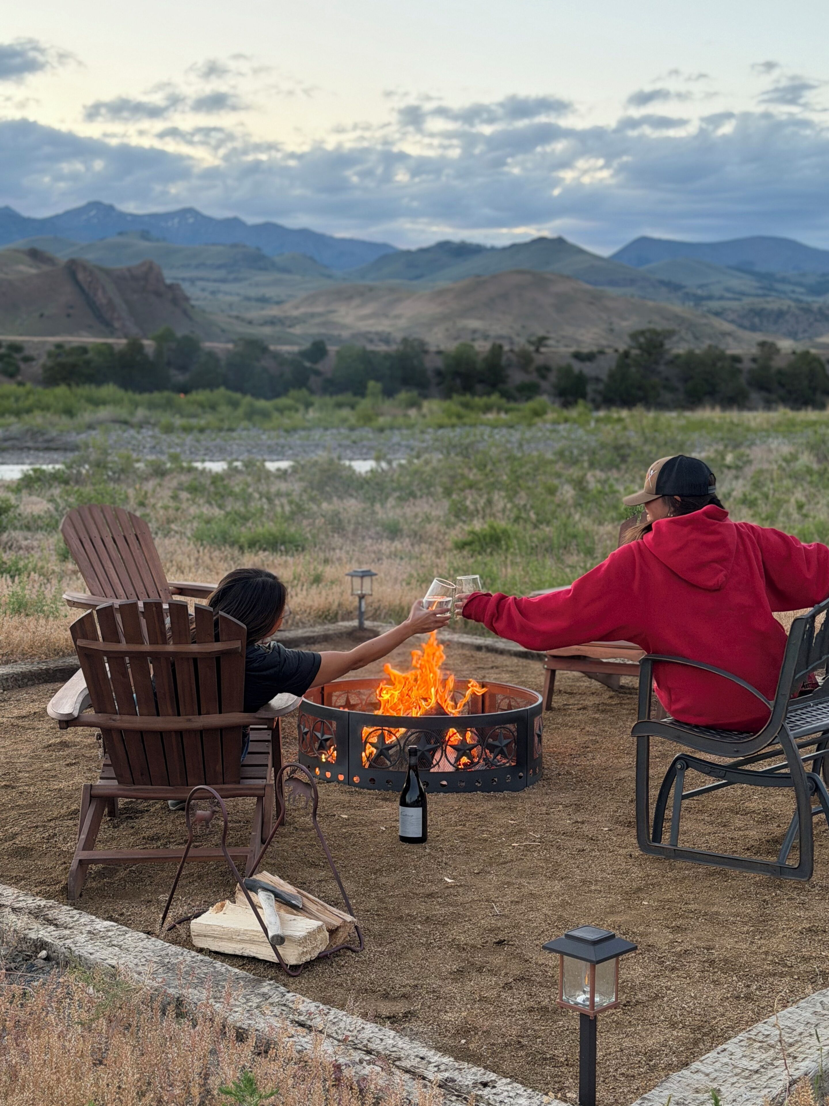 Two people toasting with drinks around a fire pit with a bottle of wine, surrounded by chairs and a lantern, set against a mountainous backdrop.