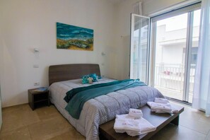 Ein Schlafzimmer mit einem Bett, einem Gemälde, einem Nachttisch, einer Bank mit Handtüchern und einem Balkon.
