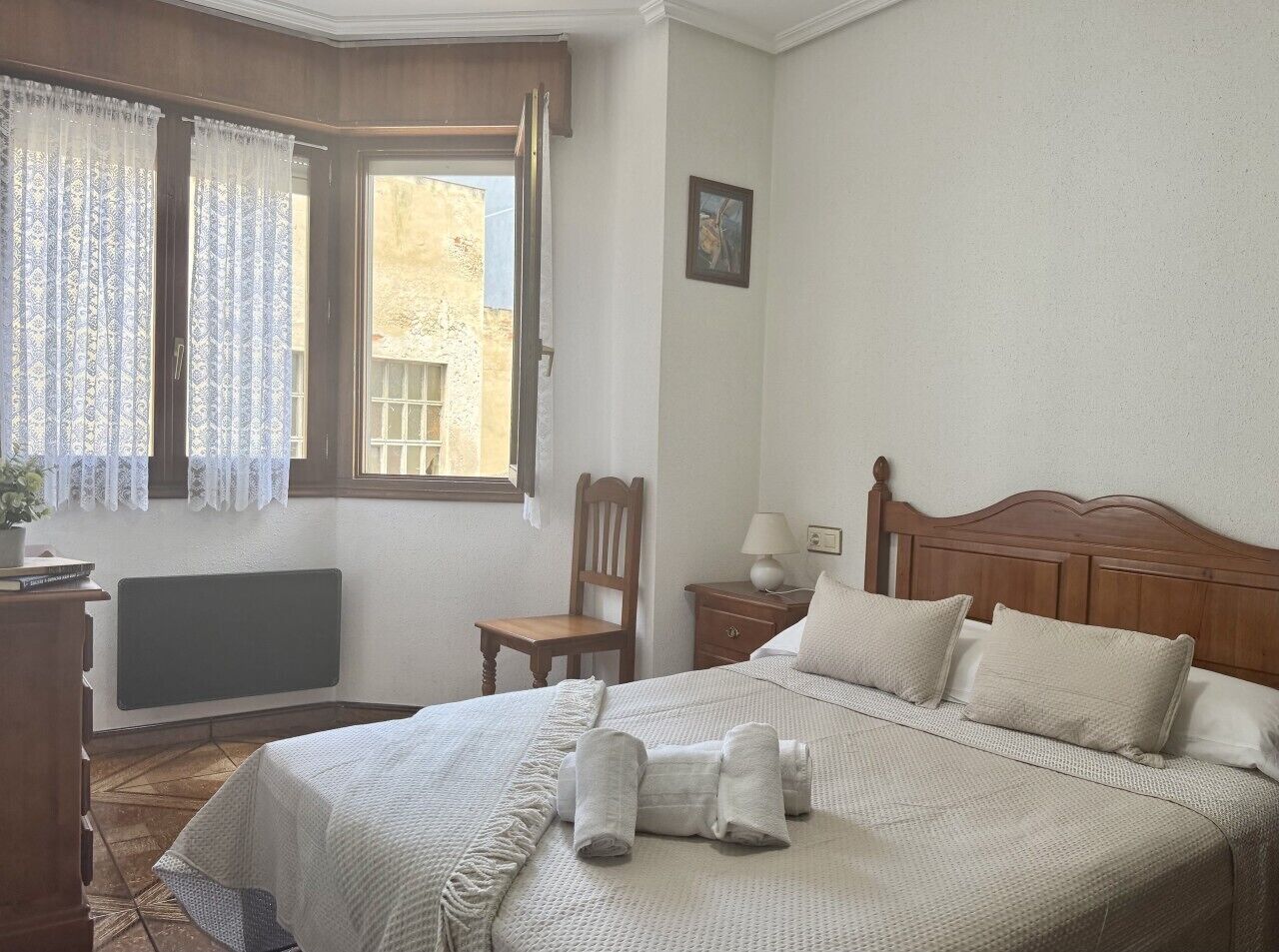 A bedroom with a bed, a chair, a nightstand, a lamp, a radiator, and a window with curtains.