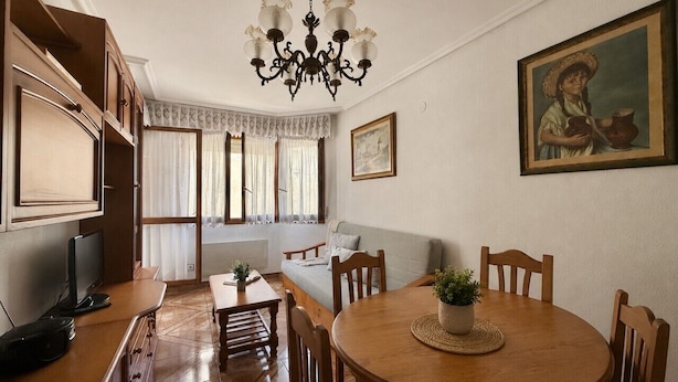 Un comedor con una mesa de madera redonda, sillas, una lámpara de araña, un sofá, un televisor y una ventana con cortinas.