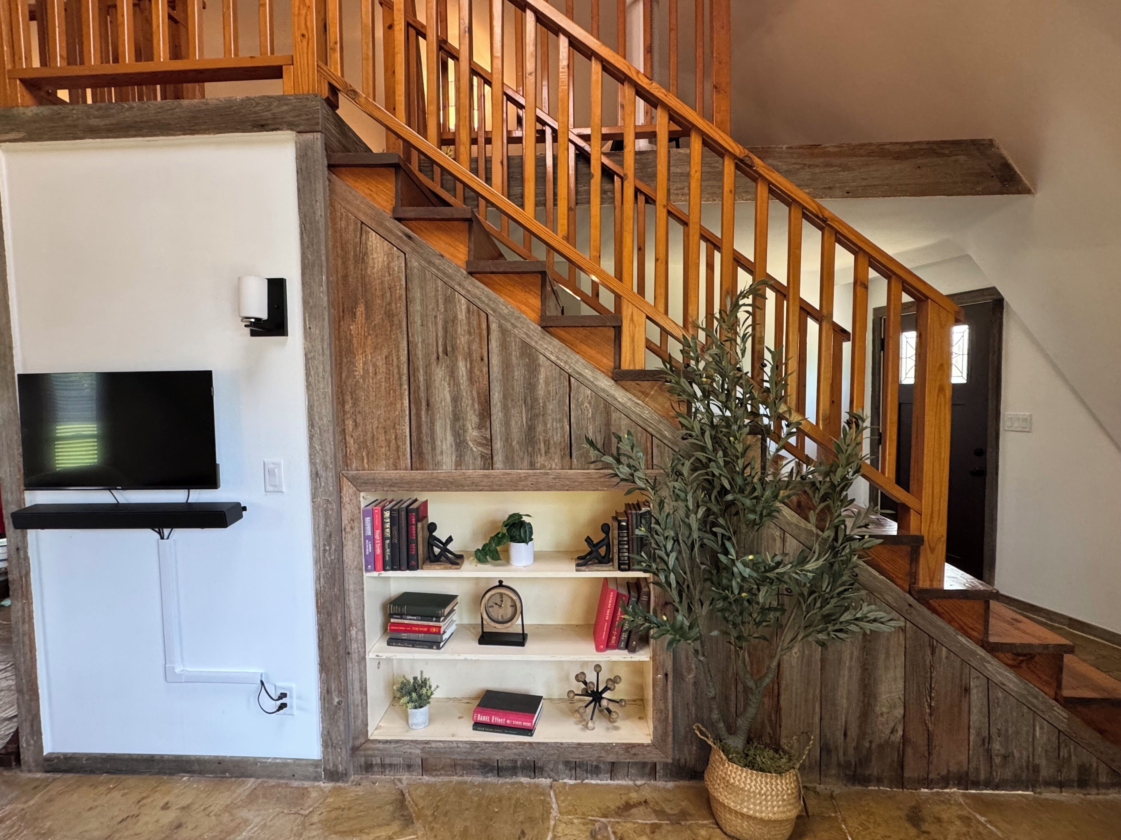Un escalier avec des rampes en bois, un téléviseur à écran plat fixé à un mur blanc, une bibliothèque remplie de livres et d’objets décoratifs, et une plante en pot.