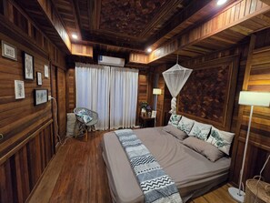 A wooden-paneled room with a bed, a chair, a bedside table with a lamp, and a window with curtains. - three little birds  2 bedroom  (Kanchanaburi)