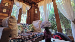 A cozy room with a wooden ceiling, a chandelier, a sofa with patterned cushions, a table with a vase and a candle, and large windows with curtains.