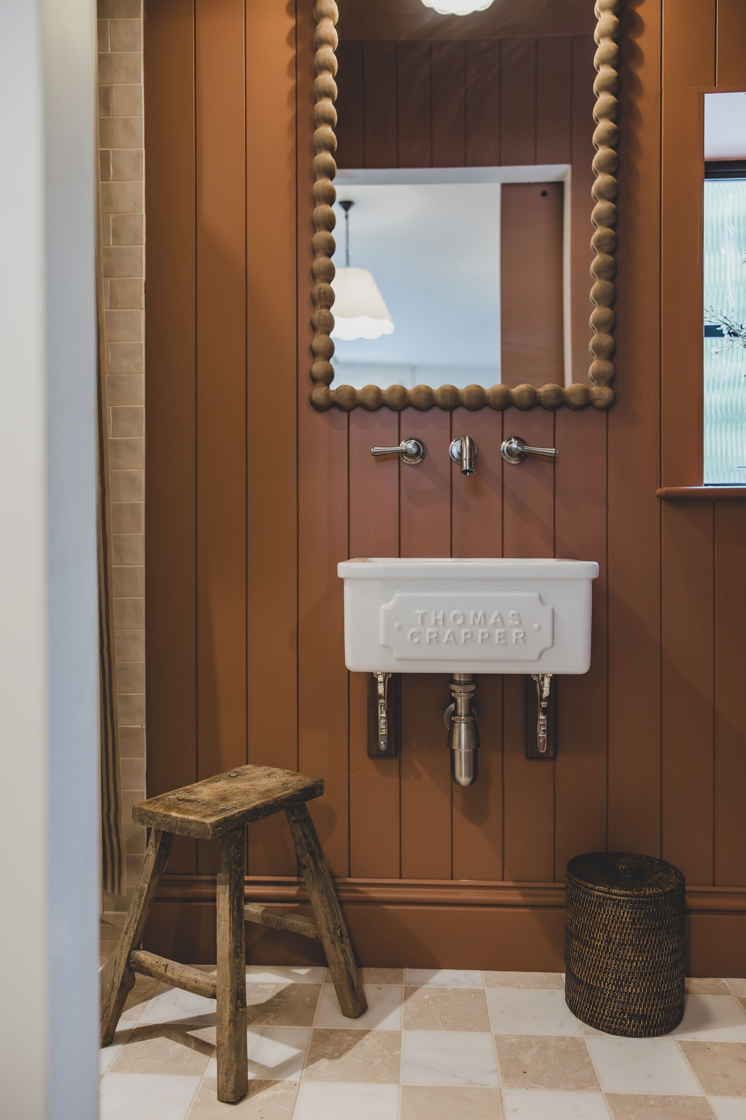 Une salle de bain avec un meuble-lavabo en bois, un lavabo blanc portant l’inscription « THOMAS CRAPPER », un miroir avec un cadre orné de perles et un petit tabouret.