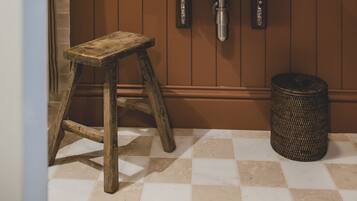 Une salle de bain avec un meuble-lavabo en bois, un lavabo blanc portant l’inscription « THOMAS CRAPPER », un miroir avec un cadre orné de perles et un petit tabouret.