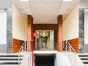 A staircase leading to a covered entrance with a metal railing, a red fire alarm, and a view of a building interior through a glass door. - Deluxe Devi Residency (Hyderabad)