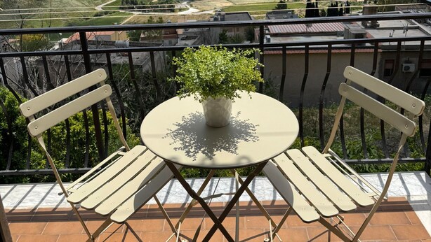 A balcony with a table and chairs, a potted plant, and a scenic view of rolling hills and a winding road.
