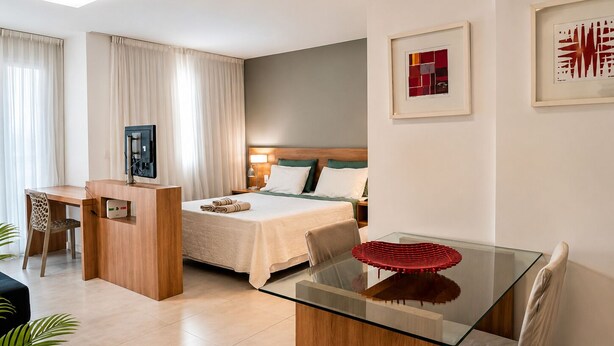 A hotel room with a bed, a desk, a chair, a TV, and artwork on the walls. - Premium Apartment | Pool, Kitchen and Parking Space (Rio de Janeiro)