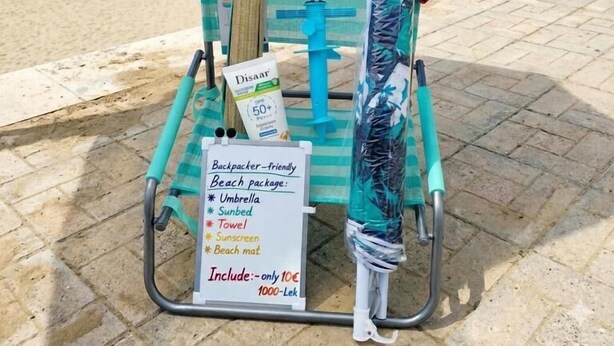 Carrito de vendedor en la playa que ofrece un paquete playero con sombrilla, tumbona, toalla, protector solar y esterilla.