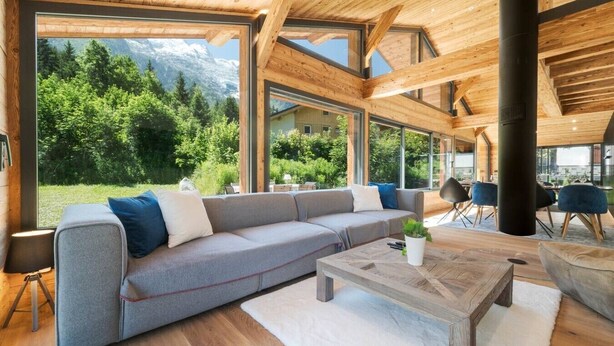 A modern interior with a large wooden ceiling, a grey sofa, and a view of a mountainous landscape. - Orion - 200 m² Chalet | 5 Bedrooms | View | Terrace & Garden (Chamonix-Mont-Blanc)