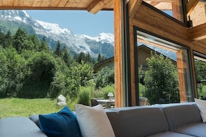 A wooden-framed room with a view of snow-capped mountains and a green landscape. - Orion - 200 m² Chalet | 5 Bedrooms | View | Terrace & Garden (Chamonix-Mont-Blanc)
