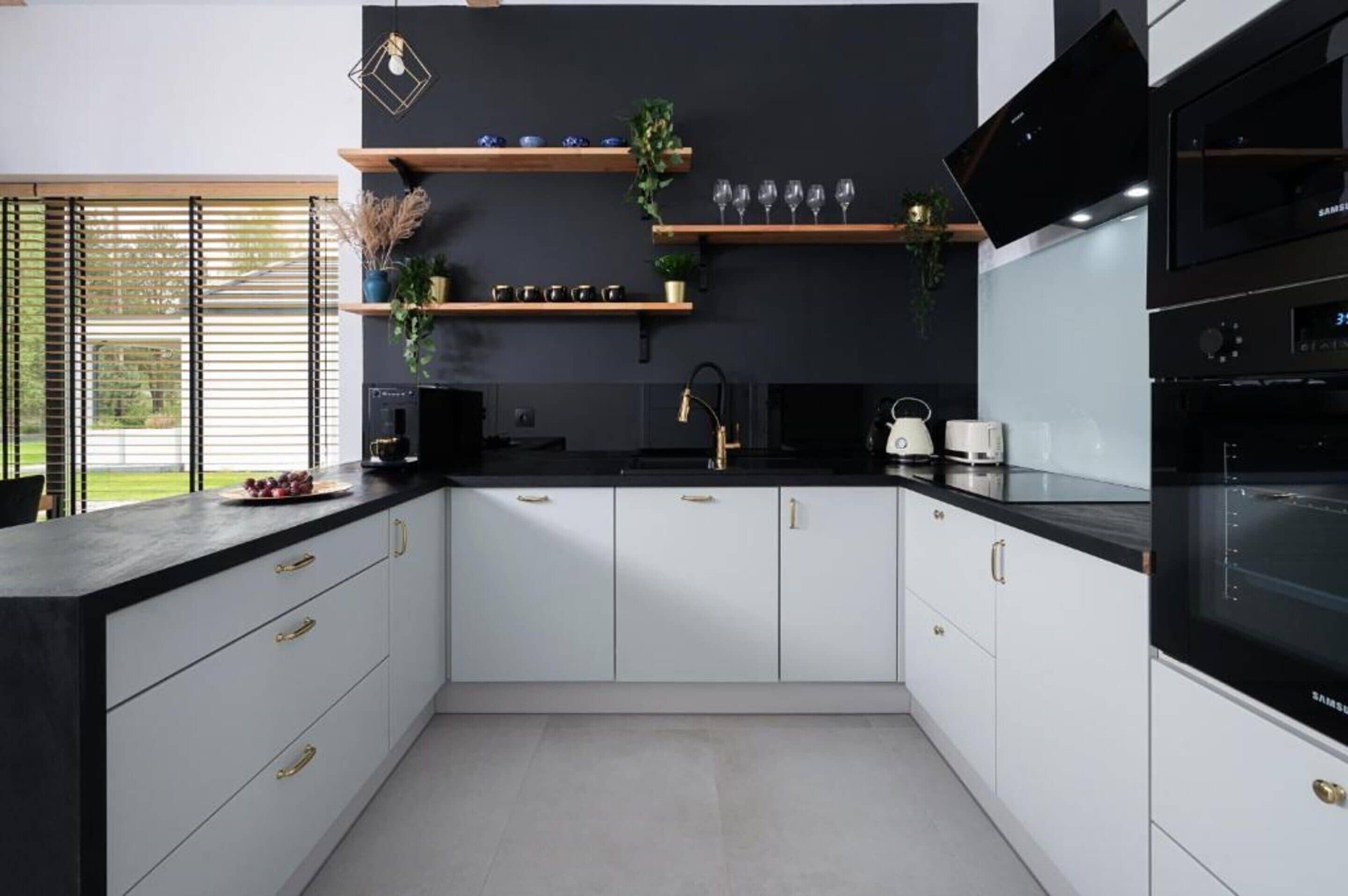 A modern kitchen with white cabinets, black countertops, and a black backsplash. There are two black ovens, a microwave, and a sink with a gold faucet.