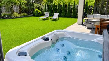 A hot tub with a grassy yard, two lounge chairs, and a wooden table.
