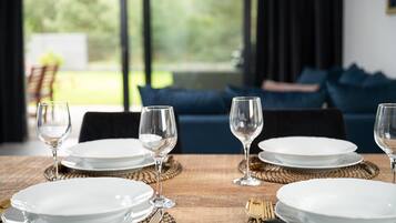 A dining table set with white plates and wine glasses, with a view of a green outdoor area through large windows.
