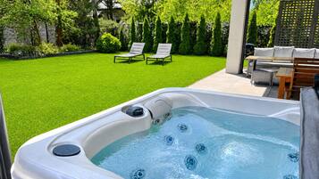 A hot tub with a patio area, two lounge chairs, and a wooden table with cushions.