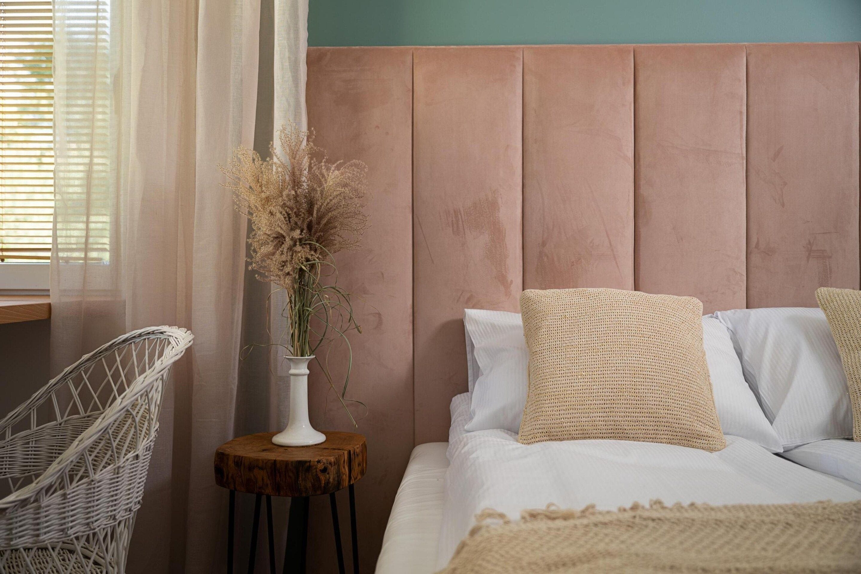 Une chambre à coucher avec un lit, une table de chevet ornée d’un vase contenant des plantes séchées, un fauteuil en osier et une fenêtre avec des stores.