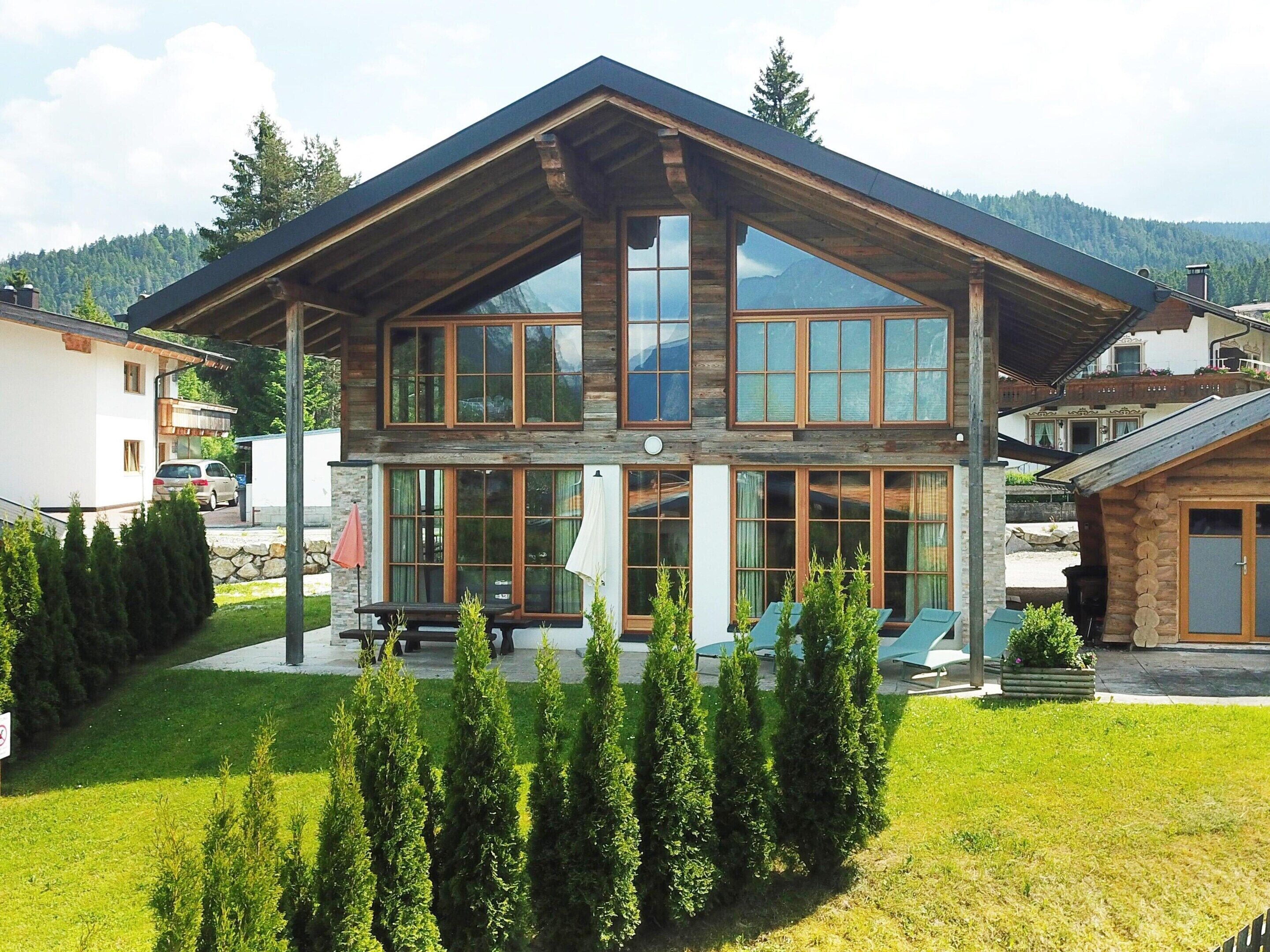 A modern two-story house with large windows, a covered patio, and a mountain backdrop.